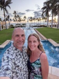 Hawaii (2021) Couple taking a selfie in a tropical resort setting with palm trees and a water feature, showcasing a beautiful sunset ambiance, reflecting the vacation planning expertise of Orange Grove Travel Company.