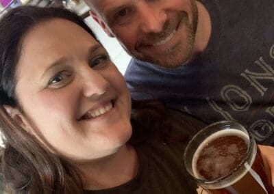 Misty Oerther and a companion smiling together, holding a glass of beer, showcasing a casual social atmosphere, reflecting personal travel experiences shared by Orange Grove Travel Company.