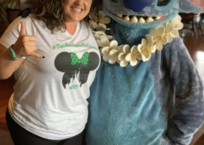 Woman in a Mickey Mouse-themed shirt posing with Stitch character at a Disney-themed event, highlighting personal travel experiences.