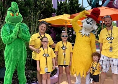 Family posing with the Grinch and Cat in the Hat characters at a theme park, wearing matching yellow shirts, surrounded by colorful umbrellas and greenery, highlighting a fun vacation experience.