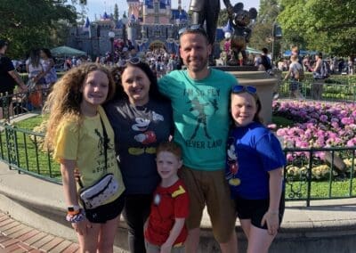 Family posing in front of Disneyland's iconic castle, wearing Disney-themed shirts, surrounded by colorful flowers and a lively park atmosphere.