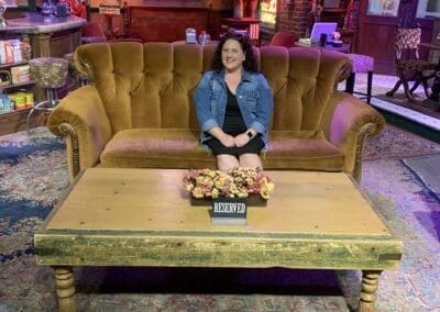 Misty Oerther sitting on a vintage couch in a themed setting, with a reserved sign on a coffee table, showcasing her travel experiences related to popular destinations.
