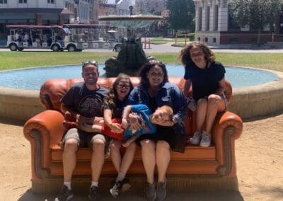 Family sitting on a large orange couch in a park, smiling together with a fountain in the background, highlighting personal travel experiences and family vacations.