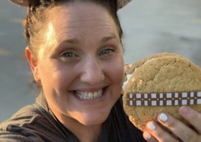 Smiling woman holding a large cookie inspired by Chewbacca, wearing mouse ears, enjoying a treat at a Disney park, reflecting personal travel experiences.