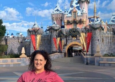 Woman in pink shirt smiling in front of Disneyland castle, showcasing personal travel experience to Disney parks.