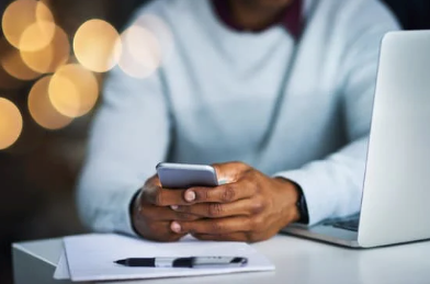 Person holding smartphone while seated at a desk with a laptop and notepad, illustrating communication for travel inquiries with Orange Grove Travel Company.