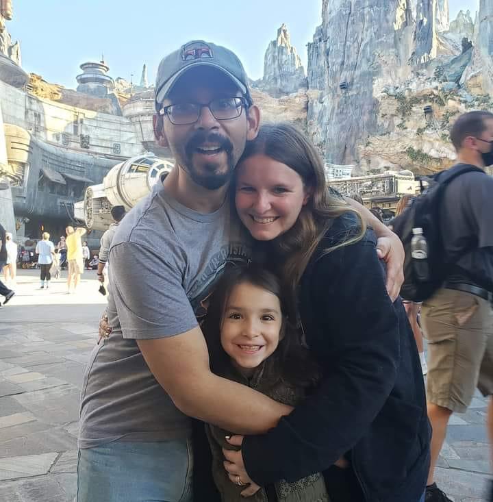Family posing together at a Disney theme park, smiling and embracing, with a Star Wars-themed backdrop featuring rocky terrain and a spaceship.