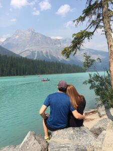 Banff Canada Couple sitting on rocks by a turquoise lake, enjoying scenic mountain view, representing relaxation and adventure in travel planning.