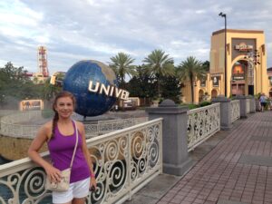 Universal Studios Orlando Woman in a purple tank top standing near the Universal Studios globe, showcasing a vibrant theme park atmosphere with palm trees and attractions in the background.