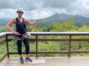 Costa Rica (2023) Woman in climbing gear gesturing towards a scenic view of Arenal Volcano in Costa Rica, showcasing adventure travel opportunities and outdoor activities.