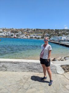 PXL_20240624_095230883 Woman standing by the shore in a coastal destination, showcasing a vibrant blue sea and white buildings in the background, reflecting the ease of vacation planning offered by Orange Grove Travel Company.
