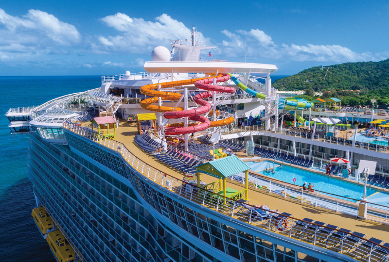 cruises_small2 Cruise ship deck featuring colorful water slides, sun loungers, and a swimming pool, set against a backdrop of ocean and blue skies, highlighting vacation experiences for travelers.